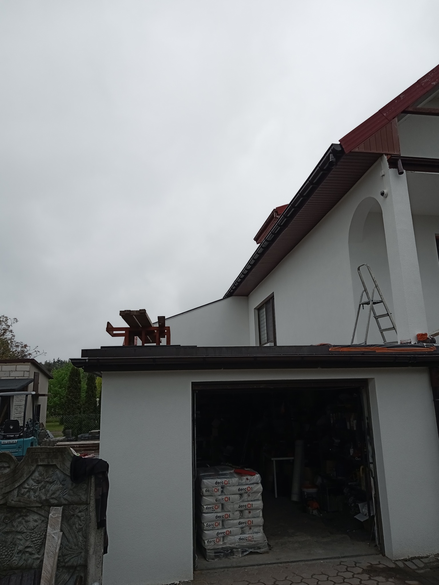 Biała elewacja domu jednorodzinnego z garażem, widoczne rusztowanie na dachu, drabina oparta o ścianę, worki z materiałem budowlanym w garażu, pochmurne niebo.