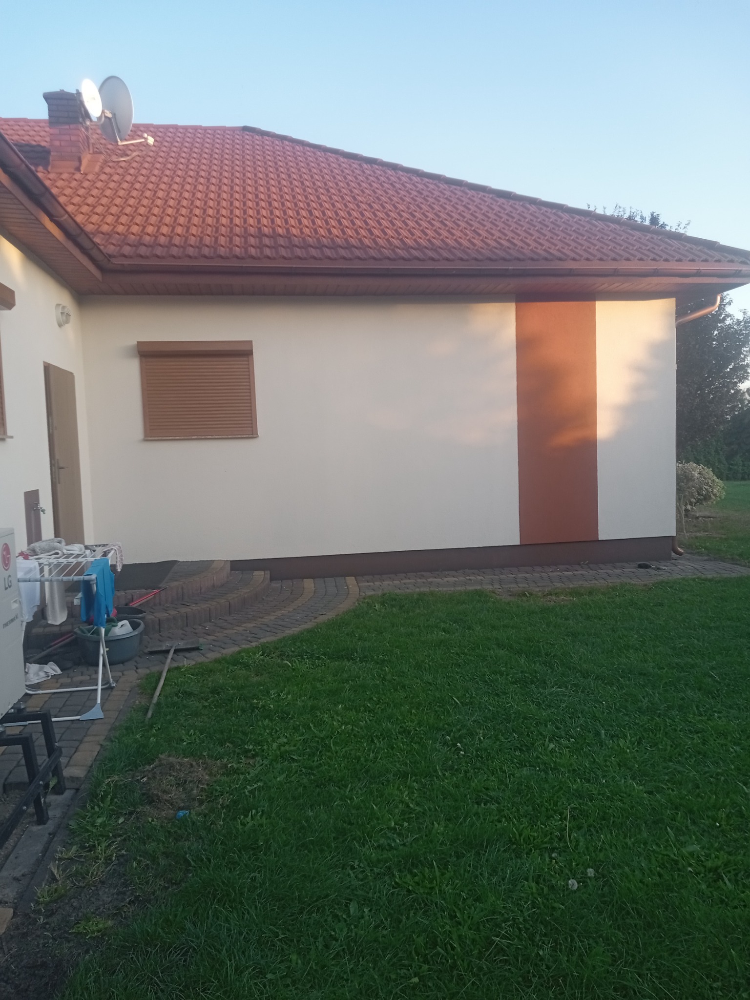 Boczny widok domu z jasną elewacją z brązowym pionowym pasem, brązową roletą okienną i czerwoną dachówką, widoczne pranie na suszarce i trawnik.