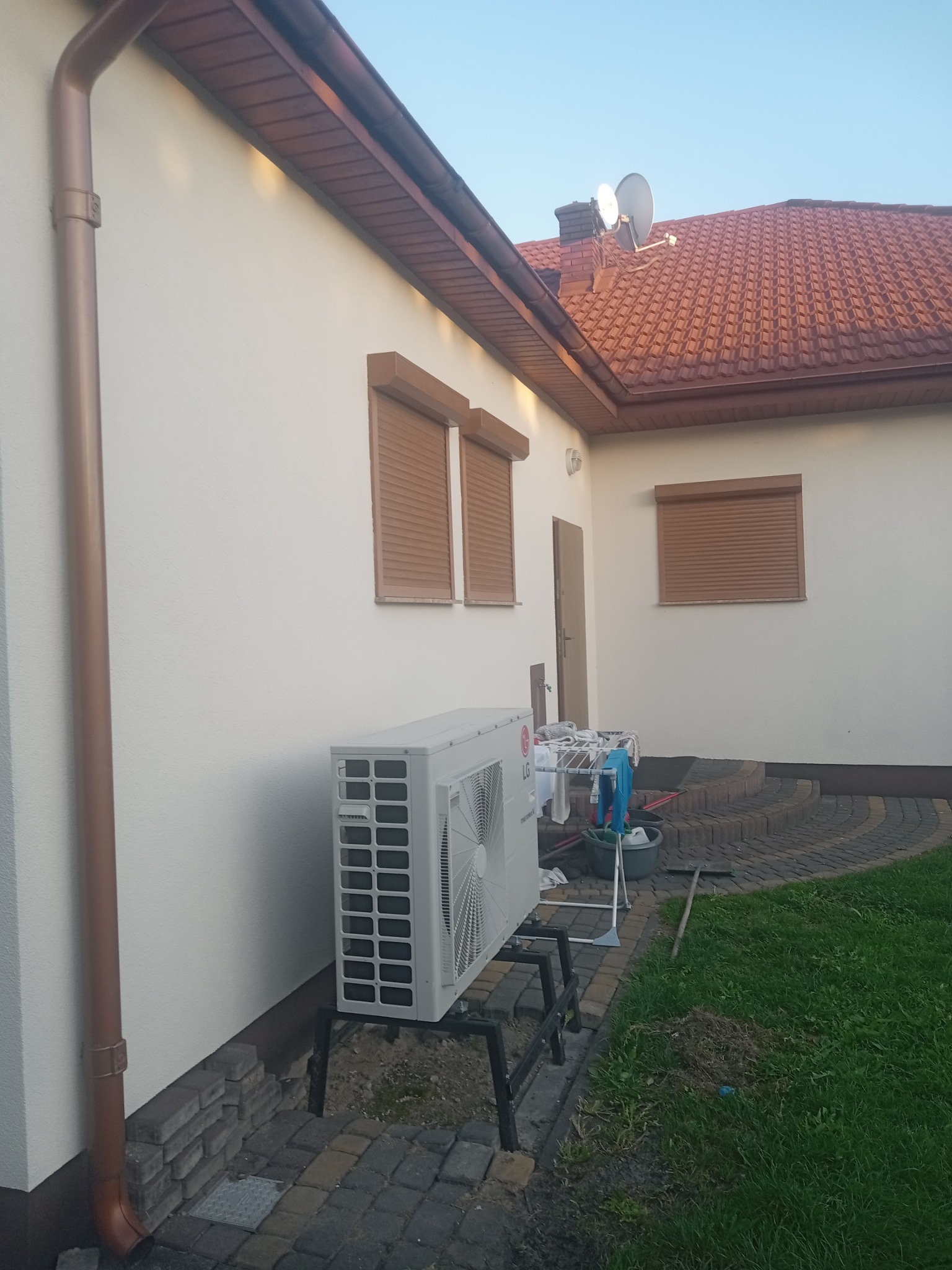Biała elewacja domu z brązowymi roletami zewnętrznymi, brązową rynną i zamontowaną jednostką zewnętrzną klimatyzacji LG na czarnym stelażu, widoczne pranie na suszarce, kostka brukowa i trawnik.