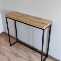 Konsola z blatem z jasnego drewna i czarną, metalową ramą w minimalistycznym stylu, ustawiona na panelach podłogowych w odcieniach brązu, na tle jasnej ściany.