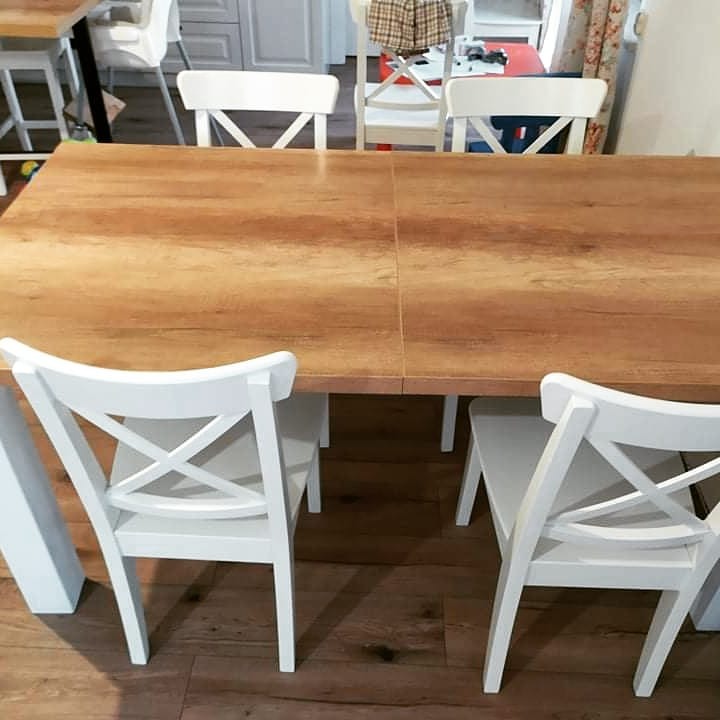 Rozkładany stół z blatem o strukturze drewna i białymi krzesłami z krzyżowym oparciem, widok z bliska na łączenie blatów.
