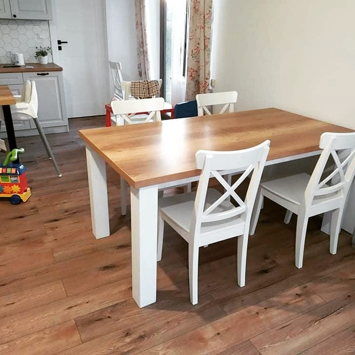 Drewniany stół jadalny z białymi nogami i krzesłami w jasnym wnętrzu, widok na kuchnię i dziecięcą zabawkę na podłodze.