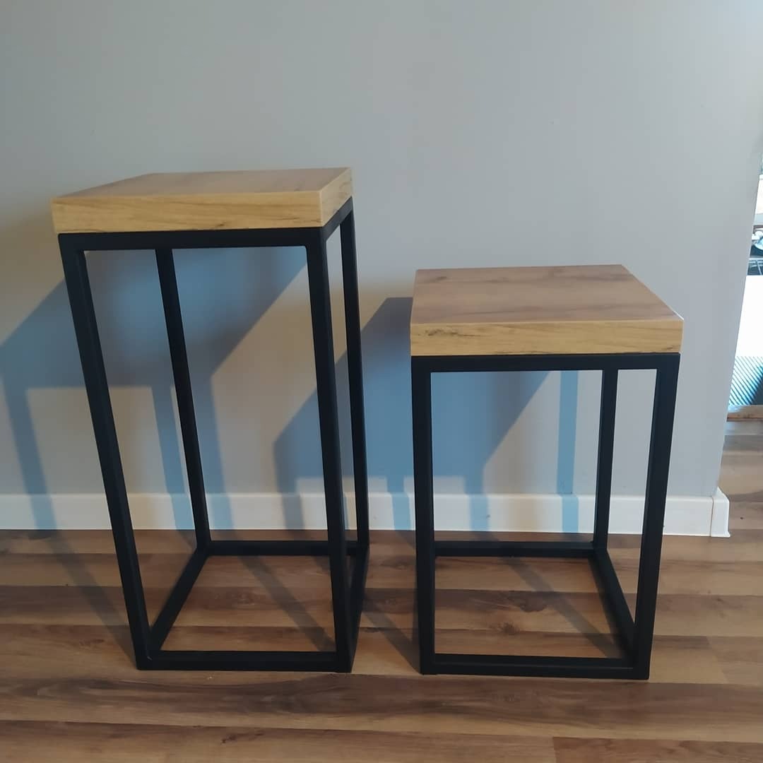 Dwa stoliki pomocnicze: wyższy z trapezoidalną podstawą i niższy z kwadratową, oba z drewnianymi blatami i czarnymi, metalowymi ramami, ustawione na podłodze imitującej drewno na tle szarej ściany.