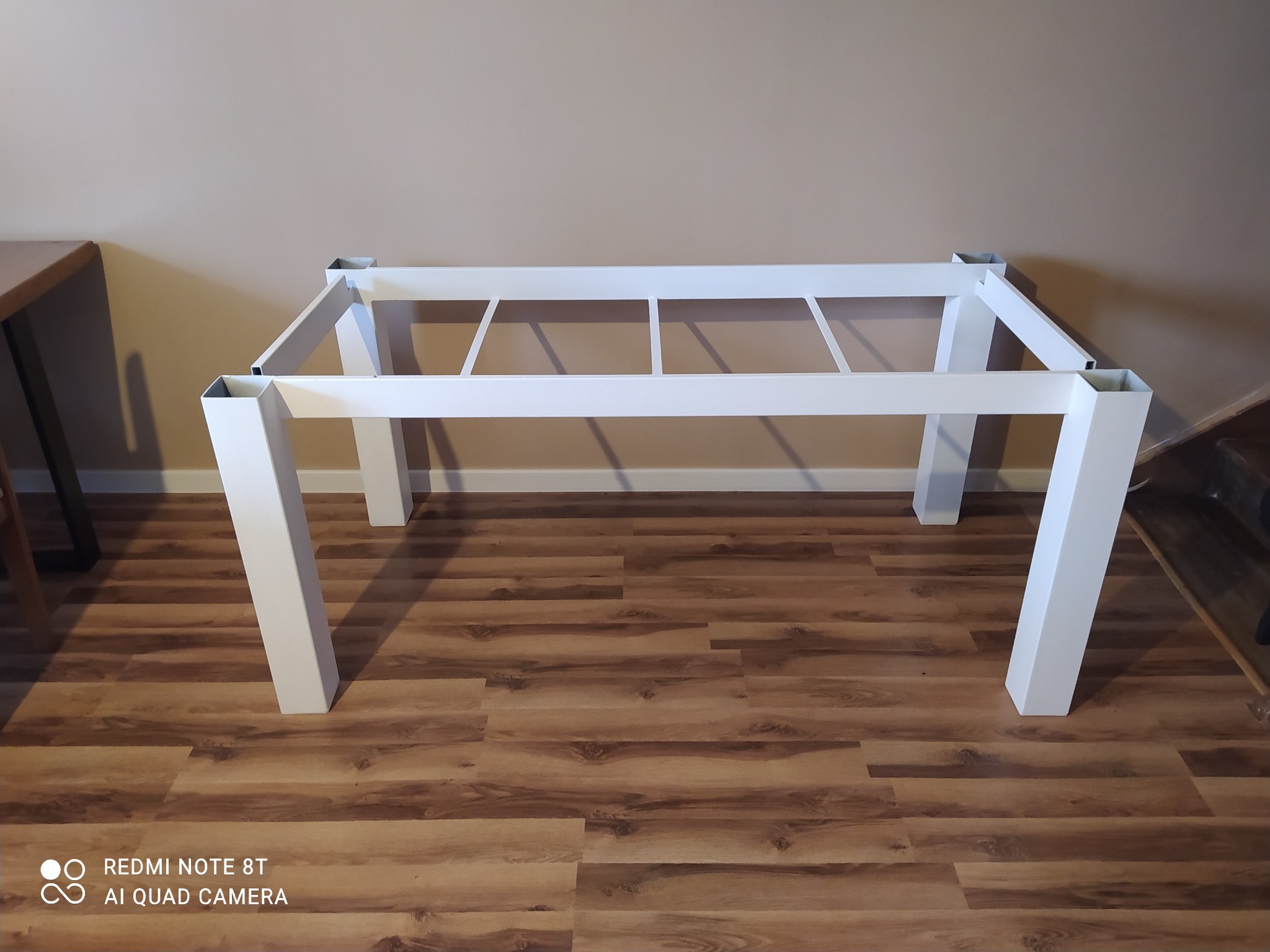 Metalowa, biała rama stołu z kwadratowymi nogami, bez blatu, na drewnianej podłodze.