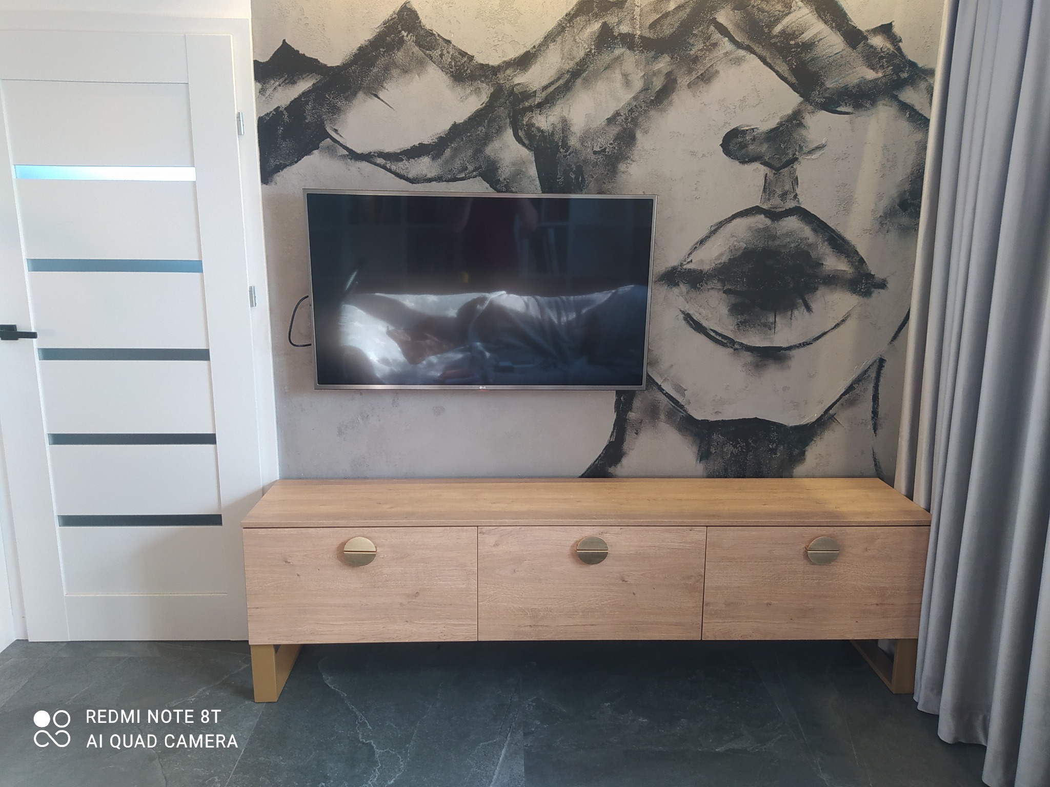 Jasna, drewniana szafka RTV z trzema szufladami o okrągłych uchwytach, ustawiona pod telewizorem na tle ściany z abstrakcyjnym malunkiem twarzy.