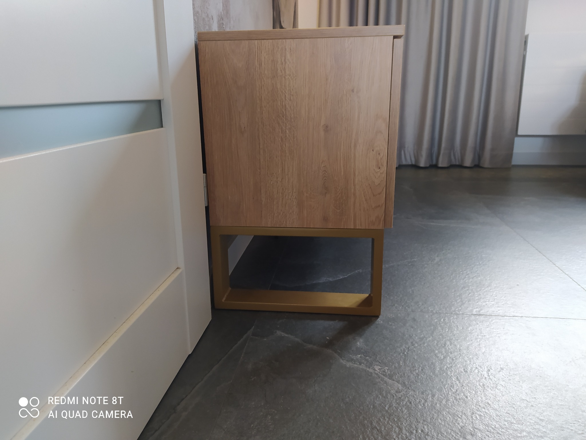 Szafka nocna z drewnianymi frontami na złotych nóżkach, stojąca obok białych drzwi na szarej podłodze z kamienia.