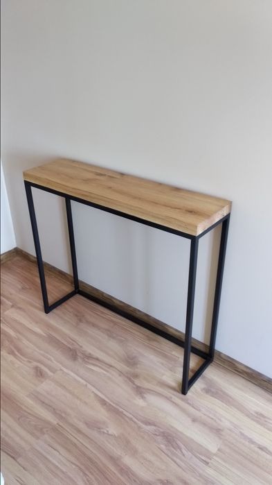 Konsola z blatem z jasnego drewna i czarną, metalową ramą w minimalistycznym stylu, ustawiona na panelach podłogowych w odcieniach brązu, na tle jasnej ściany.