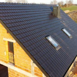 Dom w stanie surowym otwartym, z brązowym dachem z dachówki ceramicznej, widoczne okna dachowe i komin, cegły na ziemi i wznoszone ściany.