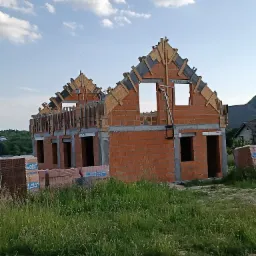 Budowa domu z czerwonej cegły, widoczne ściany w trakcie murowania, elementy drewniane konstrukcji dachu i palety cegieł Leier na pierwszym planie, trawa i błękitne niebo w tle.