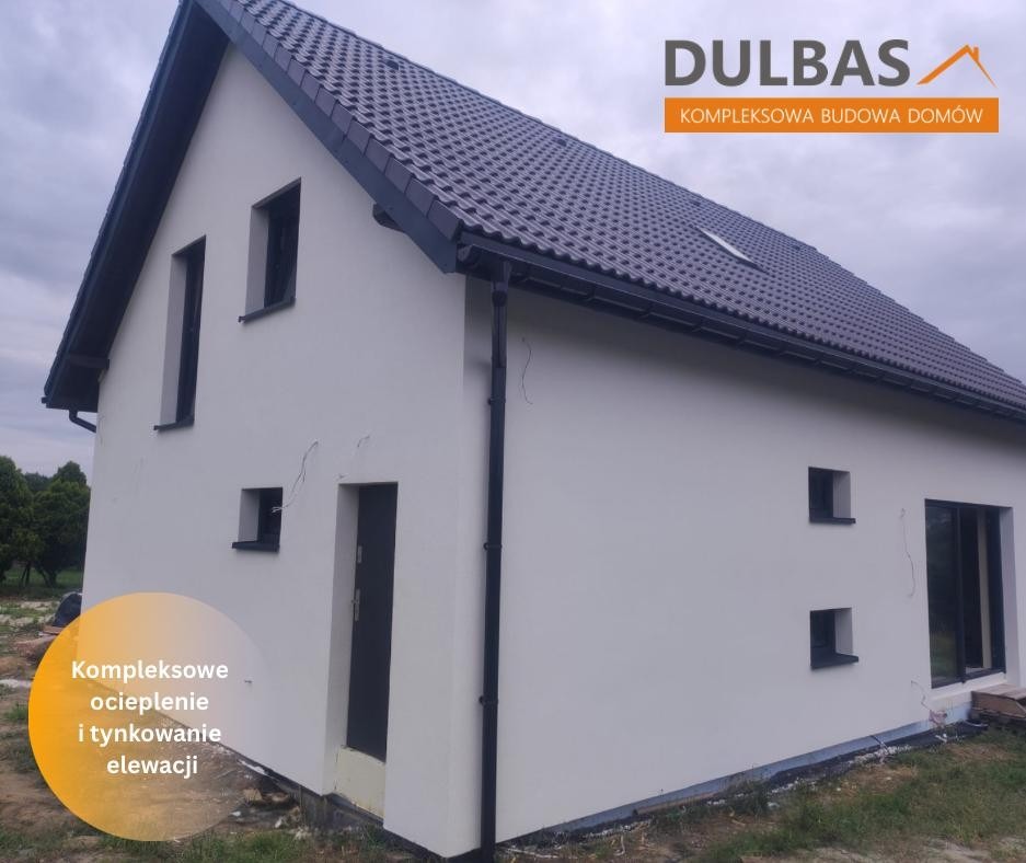 Biały dom z ciemną dachówką i orynnowaniem, widoczne pęknięcia na elewacji. Logo firmy budowlanej w prawym górnym rogu. Ujęcie z dołu, pochmurne niebo.