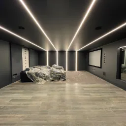 Nowoczesne kino domowe w trakcie prac wykończeniowych: czarne ściany i sufit z liniowym oświetleniem LED, jasna podłoga imitująca drewno, meble zabezpieczone folią ochronną.