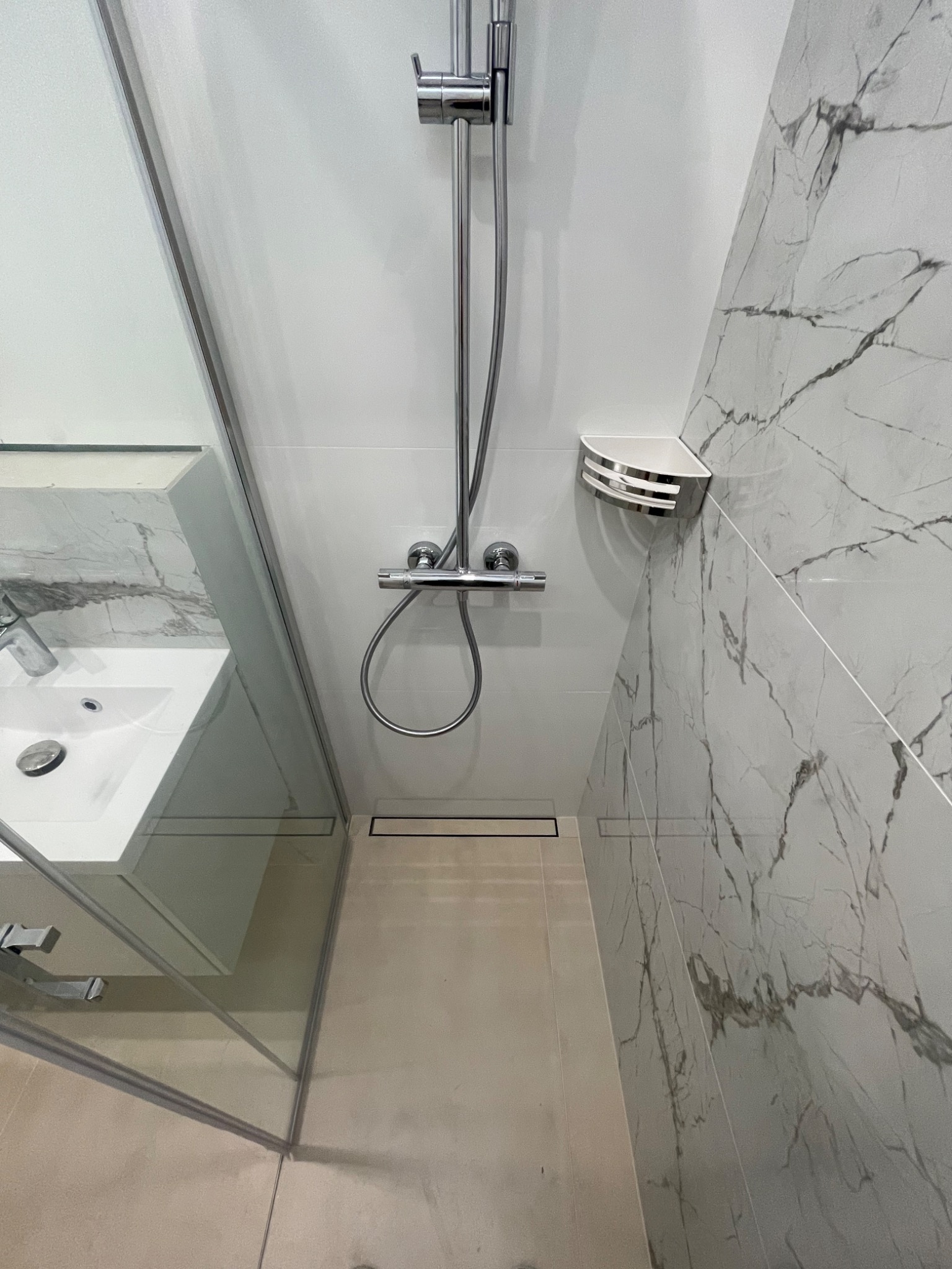 Nowoczesna kabina prysznicowa z odpływem liniowym, szklaną ścianką i baterią termostatyczną, wykończona płytkami imitującymi marmur.