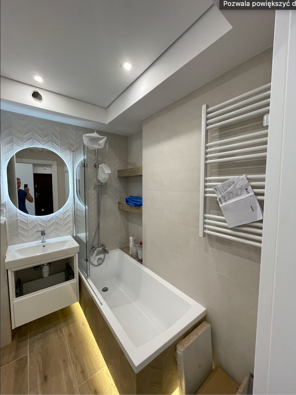 Wykończona łazienka w Rzeszowie: biała wanna z parawanem nawannowym, umywalka z szafką, grzejnik drabinkowy na ścianie. Podświetlenie LED pod wanną i szafką umywalkową.