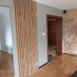 Drewniane lamele dekoracyjne na ścianie w szarym pokoju z parkietem w jodełkę, widoczny fragment ściany z efektem kamienia i otwarte drzwi do kolejnego pomieszczenia.