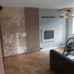 Wykończony salon z dekoracyjną ścianą z okładziną imitującą onyks, drewnianymi lamelami i obudowanym kominkiem, parkiet ułożony w jodełkę.