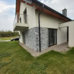 Wykończenie elewacji domu szarą cegłą klinkierową, widok z poziomu trawnika, na tle zaparkowany samochód.