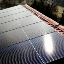 Panele słoneczne zamontowane na czerwonym dachu z dachówki ceramicznej, widoczny krajobraz z drzewami w tle, odbicie słońca na panelach.