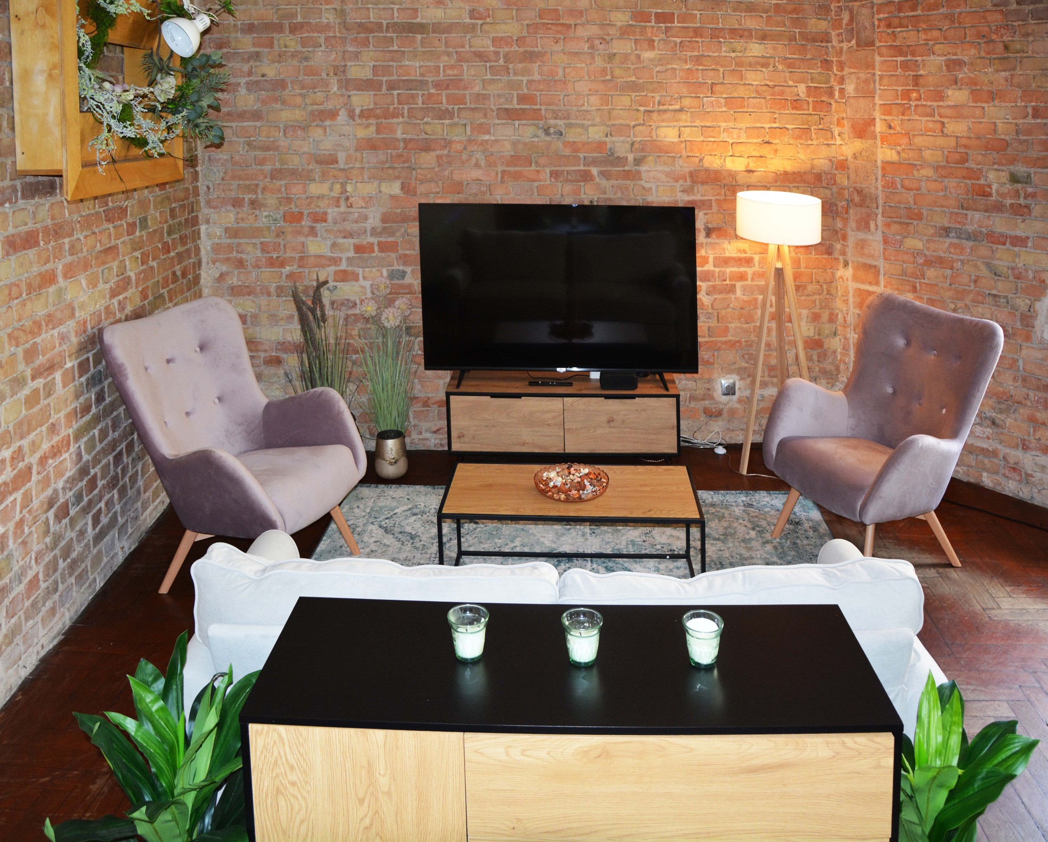Loftowy salon z ceglaną ścianą, dwoma fotelami w kolorze pudrowego różu, telewizorem na drewnianej komodzie, białym narożnikiem i trzema świecami w szklanych pojemnikach na niskiej szafce.