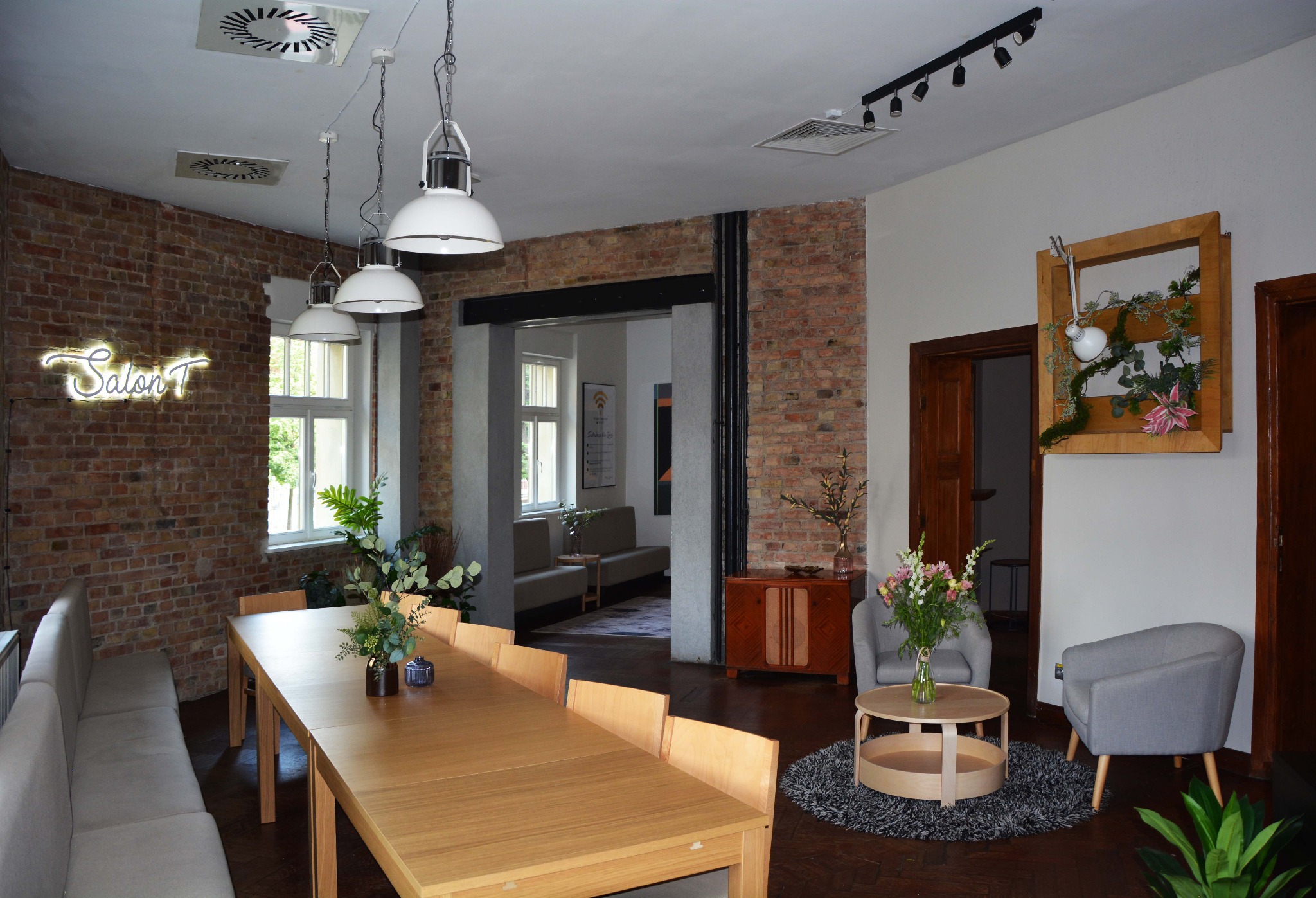 Jasne, przestronne wnętrze z długim drewnianym stołem, szarą sofą i krzesłami, dekoracjami roślinnymi, neonowym napisem 'Salon T' na ceglanej ścianie i designerskim oświetleniem.