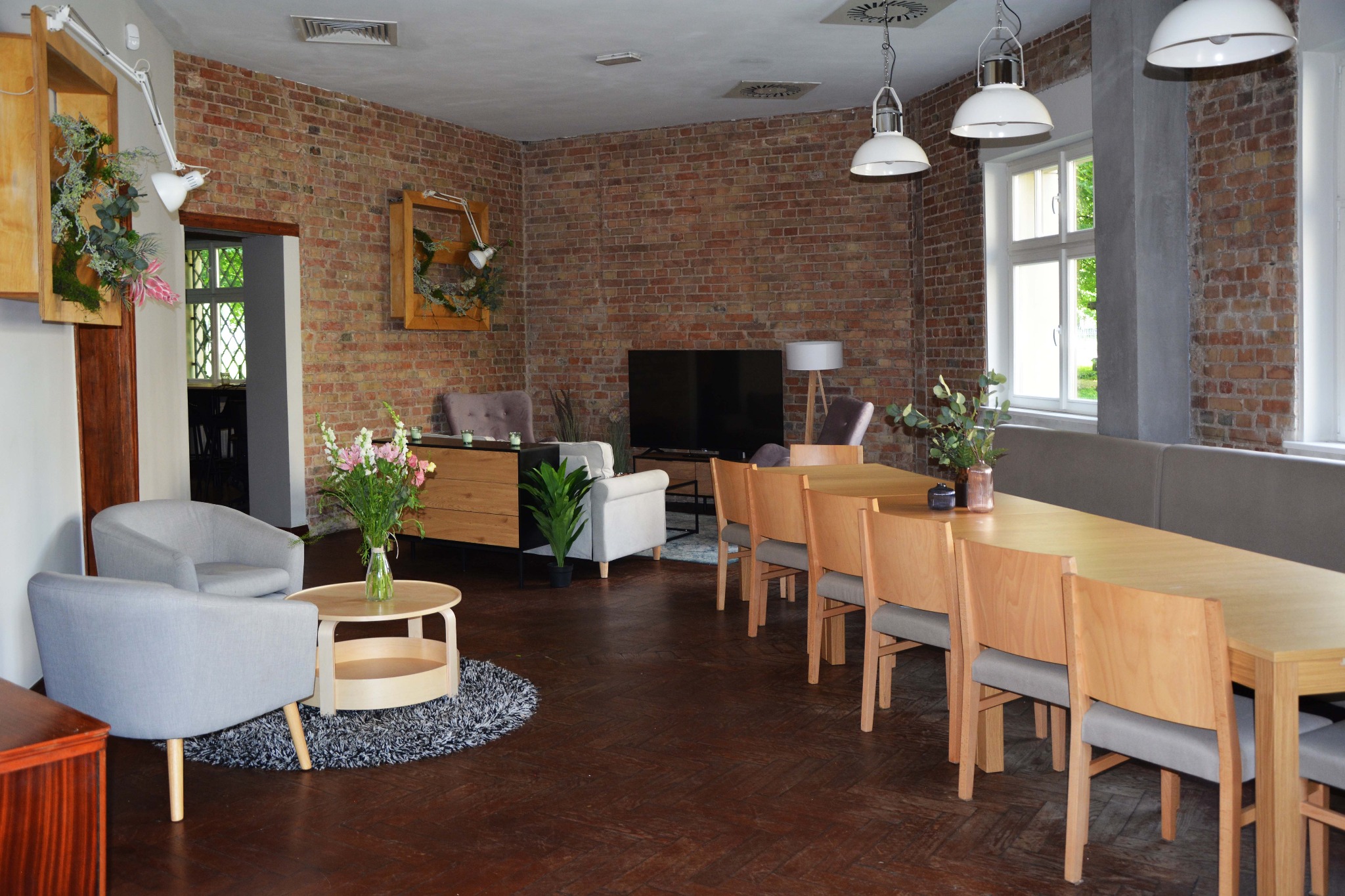 Przestronne wnętrze z ceglaną ścianą, drewnianymi stołami i krzesłami, szarymi fotelami, kwiatami w wazonach, dekoracjami ściennymi z lampkami i wiszącymi lampami w stylu industrialnym.