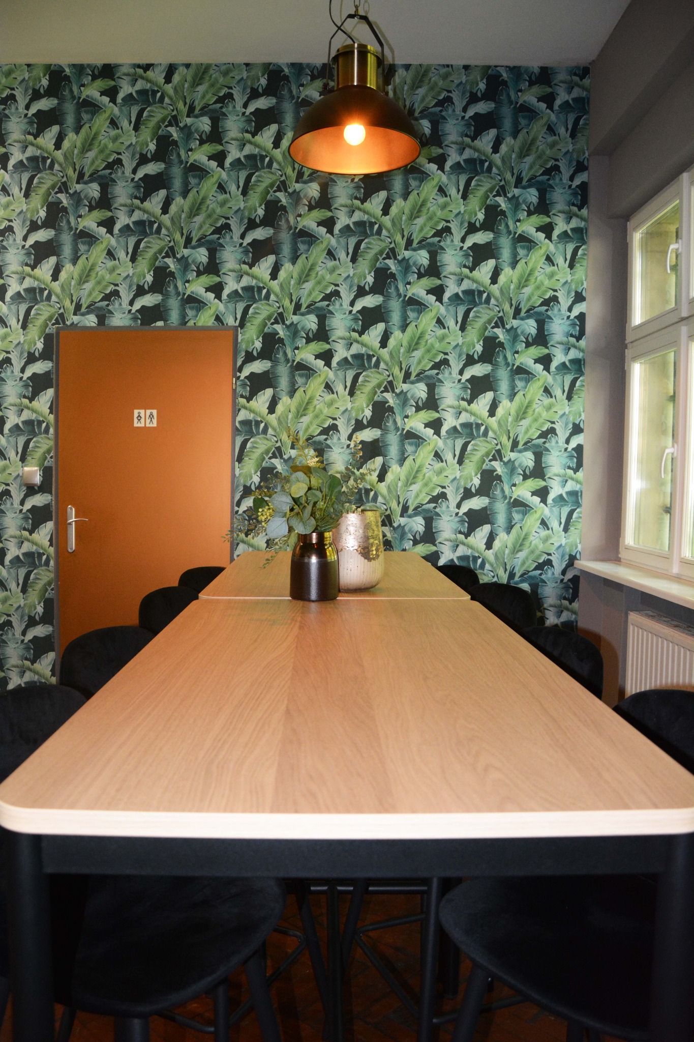 Sala konferencyjna z długim drewnianym stołem, czarnymi krzesłami i ścianą w tropikalne liście. Na stole stoją dwie dekoracyjne wazy z roślinami. Nad stołem wisi lampa w stylu industrialnym.