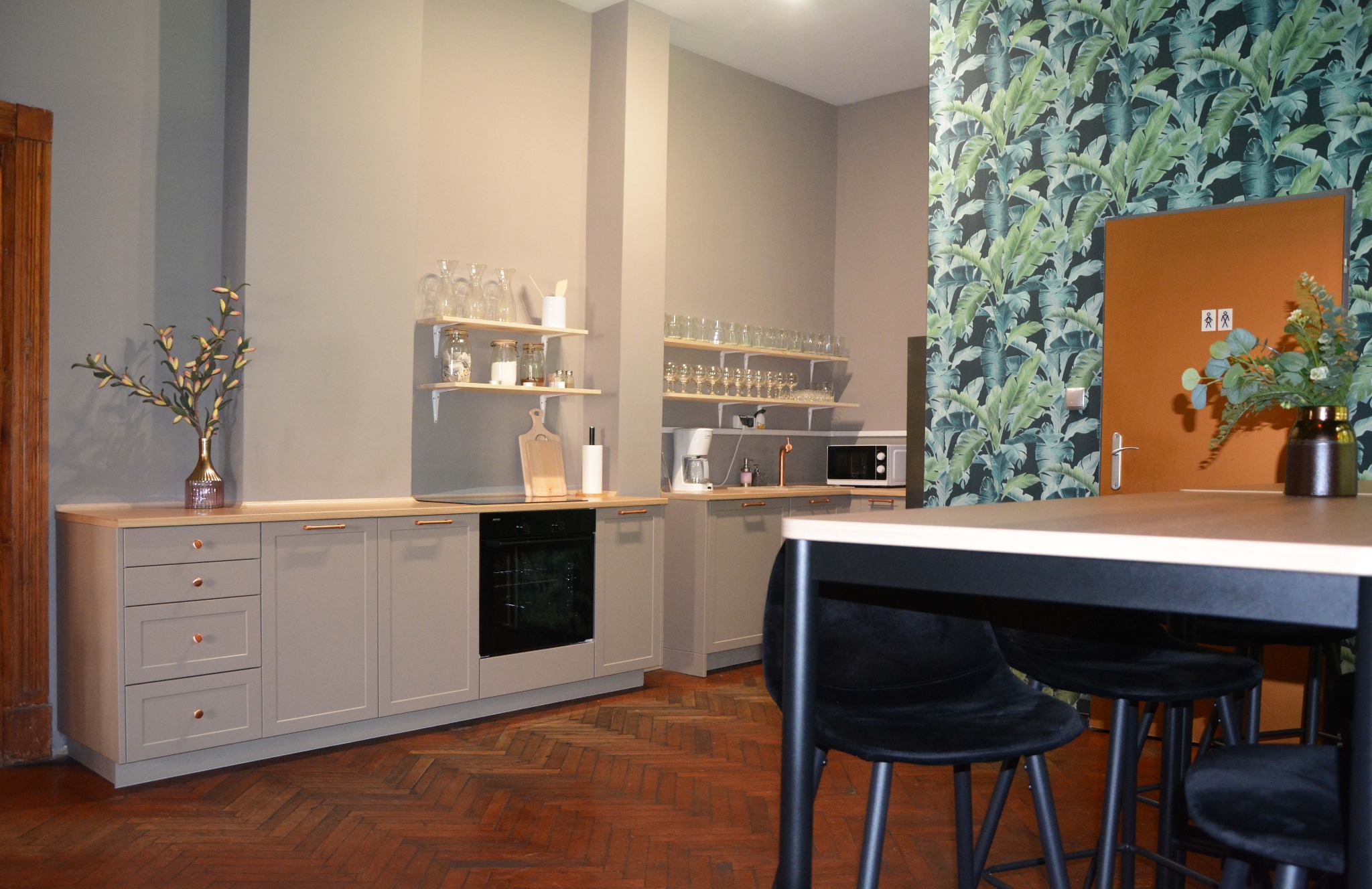 Nowoczesna kuchnia z szarymi szafkami, drewnianymi blatami i miedzianymi uchwytami, dekorowana kwiatami w wazonie i egzotyczną tapetą w liście bananowca, na podłodze parkiet w jodełkę.