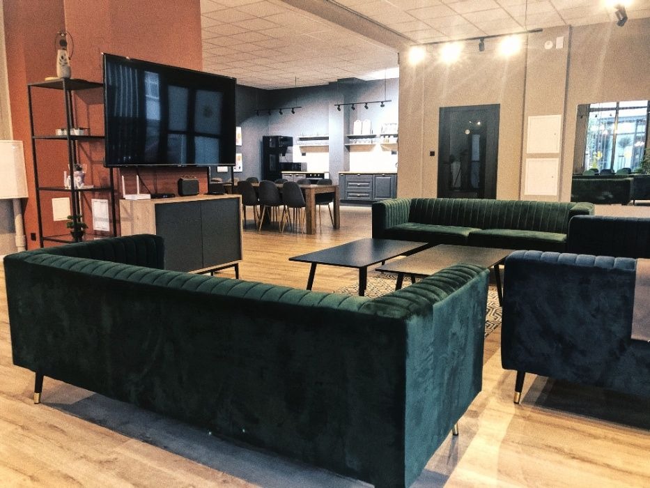 Wnętrze przestronnego salonu z drewnianą podłogą, wyposażonego w ciemnozielone, welurowe sofy o pikowanym wykończeniu, czarne stoliki kawowe oraz telewizor na szafce RTV. W tle widoczna kuchnia...