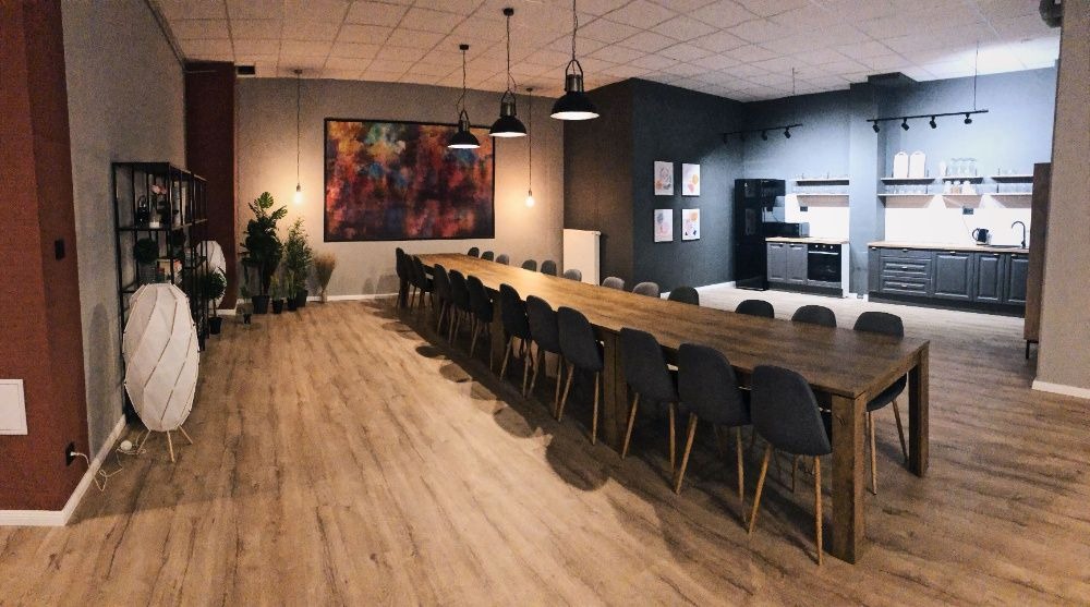 Przestronna sala konferencyjna w Poznaniu z długim drewnianym stołem otoczonym krzesłami, aneksem kuchennym w tle i abstrakcyjnym obrazem na ścianie.