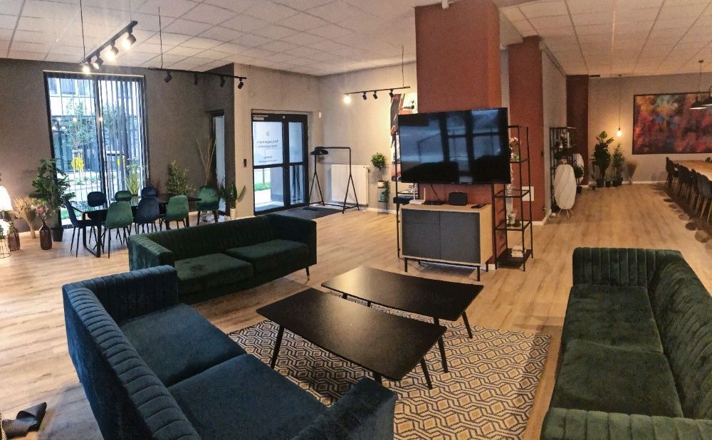 Przestronne wnętrze sali konferencyjnej lub eventowej z nowoczesnymi sofami, stolikami kawowymi, stołem konferencyjnym i telewizorem, oświetlone reflektorami i lampami.