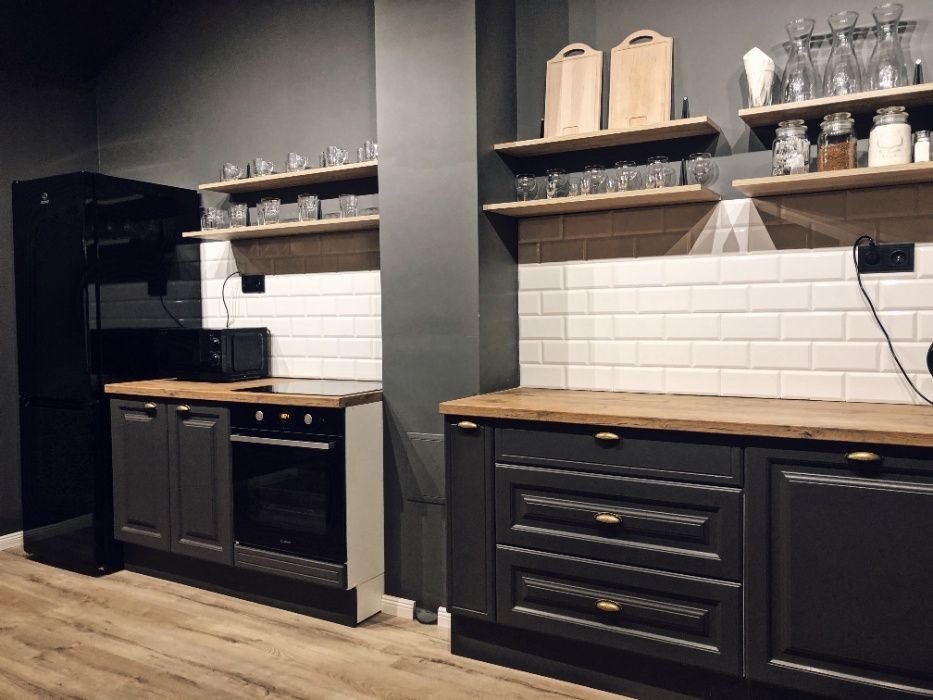 Ciemna kuchnia z drewnianymi blatami i białymi płytkami na ścianie, wyposażona w czarną lodówkę, piekarnik i mikrofalówkę, na półkach szklanki i deski do krojenia.