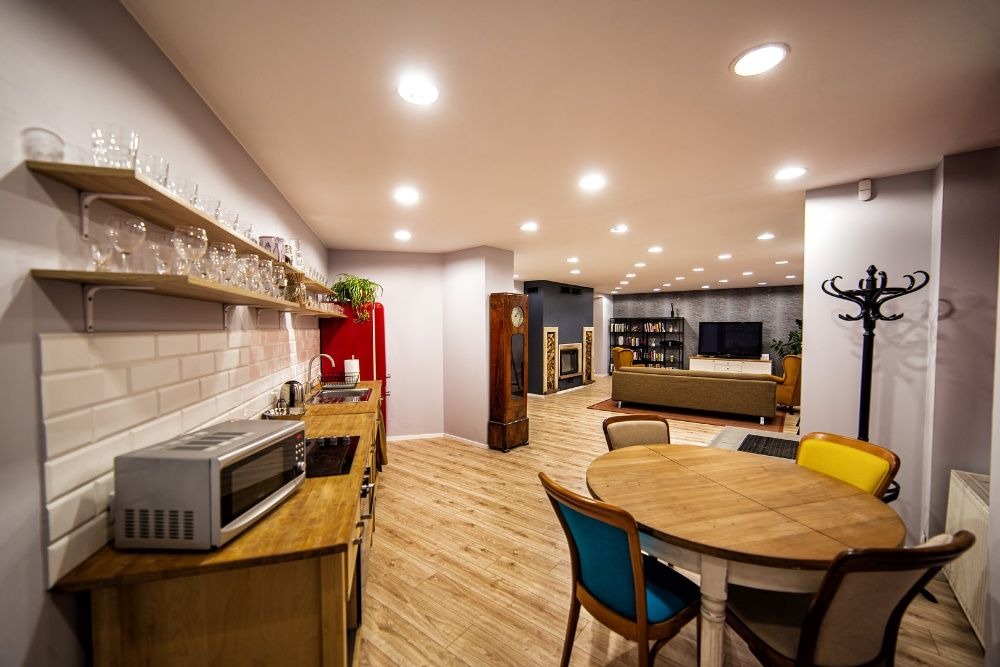 Przestronne wnętrze domu z otwartą kuchnią, jadalnią i salonem w tle. W kuchni drewniane blaty, czerwona lodówka w stylu retro i półki z kieliszkami. W jadalni okrągły drewniany stół z różnymi...