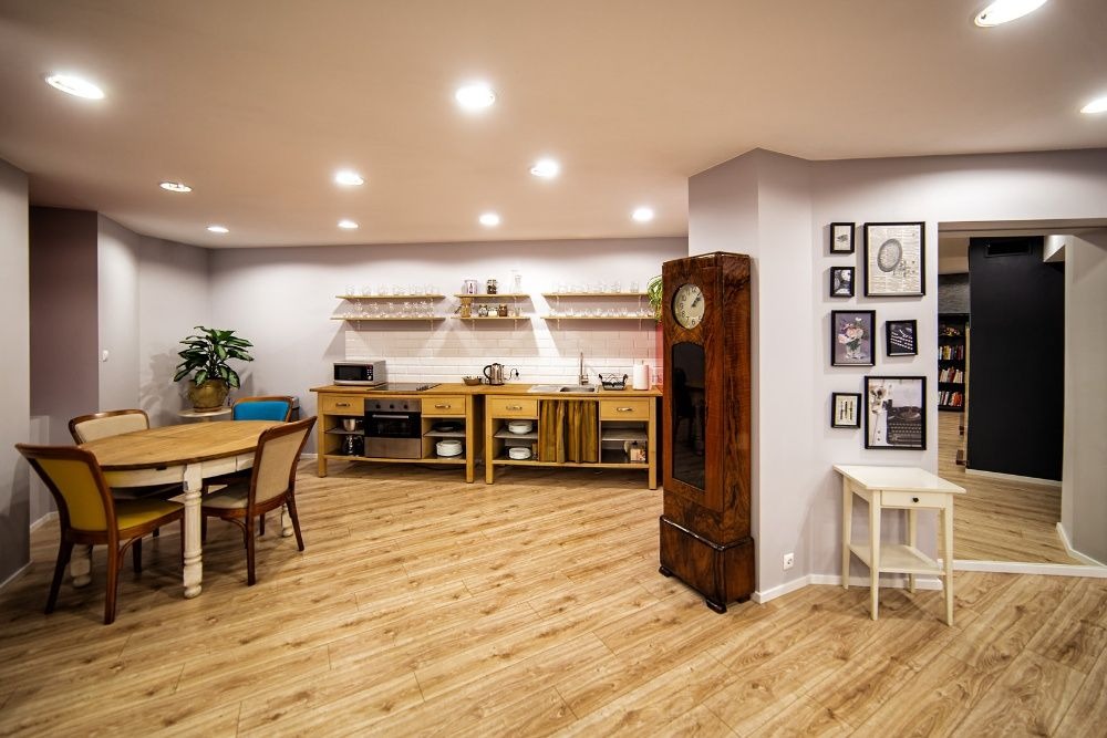 Przestronne wnętrze z drewnianą podłogą, kuchnią z drewnianymi szafkami, okrągłym stołem z krzesłami, wysokim zegarem i galerią obrazków na ścianie.