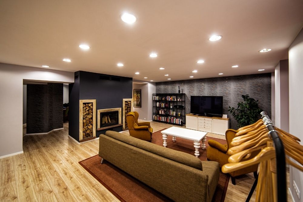 Przestronny salon z kominkiem obudowanym drewnem, brązową sofą, fotelami w kolorze musztardowym, regałem z książkami i telewizorem na tle ściany o fakturze kamienia, oświetlony punktowymi lampami...