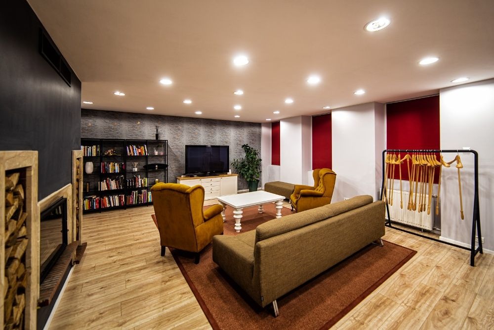 Jasne wnętrze pokoju z drewnianą podłogą, zawierające sofę, dwa fotele, stolik kawowy, regał z książkami, telewizor, kominek z drewnem i wieszak na ubrania z drewnianymi wieszakami.