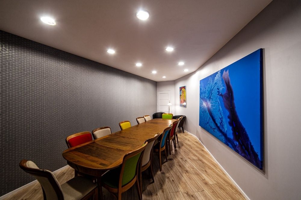 Sala konferencyjna z długim drewnianym stołem otoczonym krzesłami w różnych kolorach, ściana z geometrycznym wzorem, duży obraz z motywem morskim i punktowe oświetlenie sufitowe.