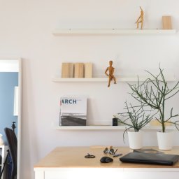 Warsztat 153 - Minimalistyczne biuro domowe z białymi ścianami, drewnianym biurkiem, półkami z dekoracjami, lustrem i czarną lampą ścienną.