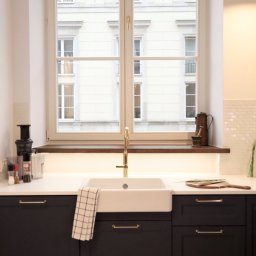 Warsztat 153 - Nowoczesna kuchnia z ciemnymi szafkami, białym blatem i ceramicznym zlewem pod oknem z widokiem na budynek.