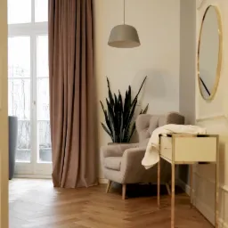 Jasny salon z parkietem ułożonym w jodełkę, beżowy fotel z rośliną, stolik z lustrem i brązowe zasłony przy oknie.