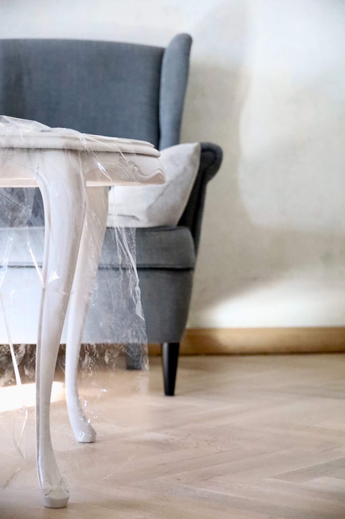Biały, odnowiony stolik z giętymi nogami zabezpieczony folią malarską, szary fotel w tle, parkiet ułożony w jodełkę.