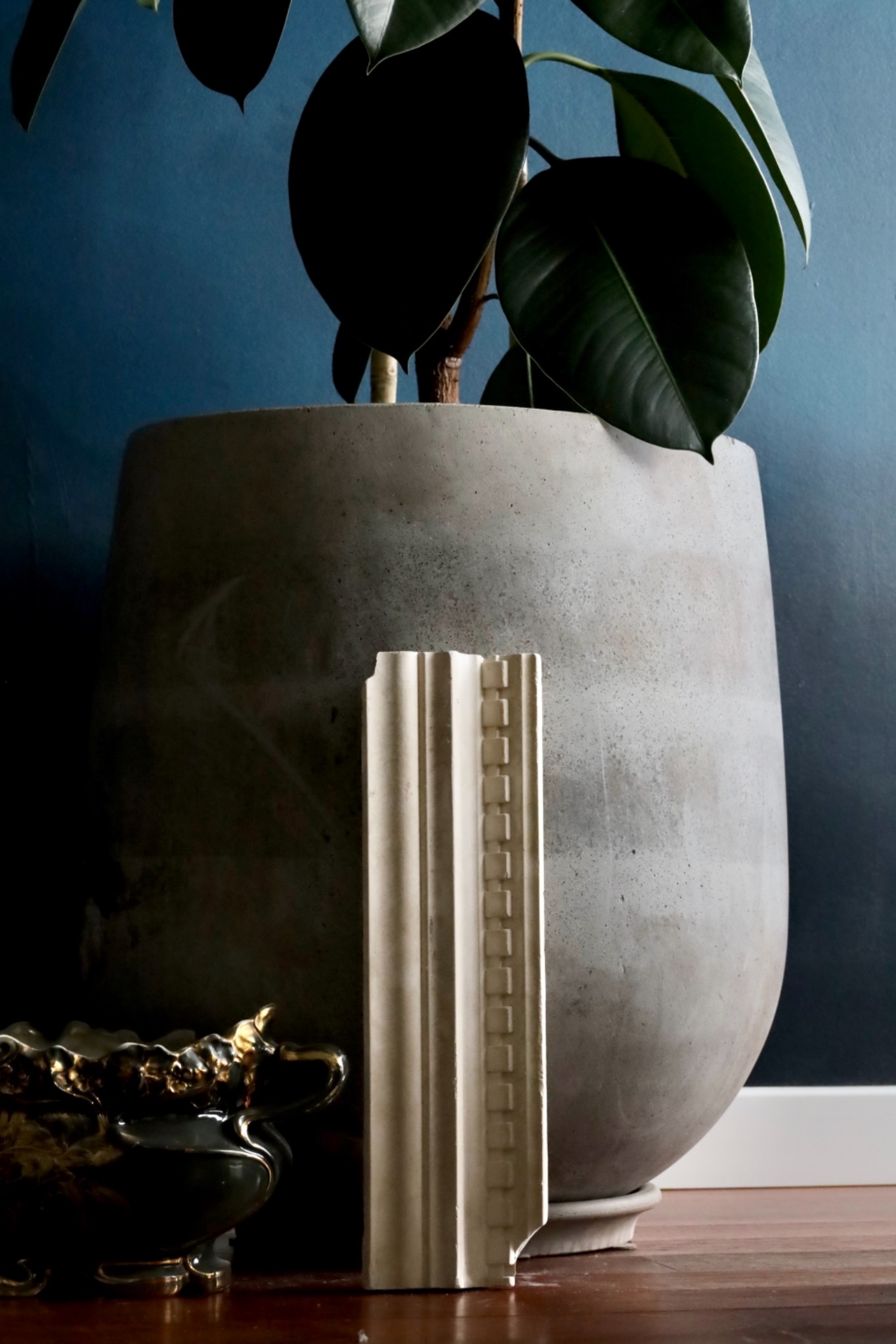 Fragment sztukaterii dekoracyjnej z gipsu, ustawiony obok donicy z rośliną na tle ściany w odcieniach granatu.