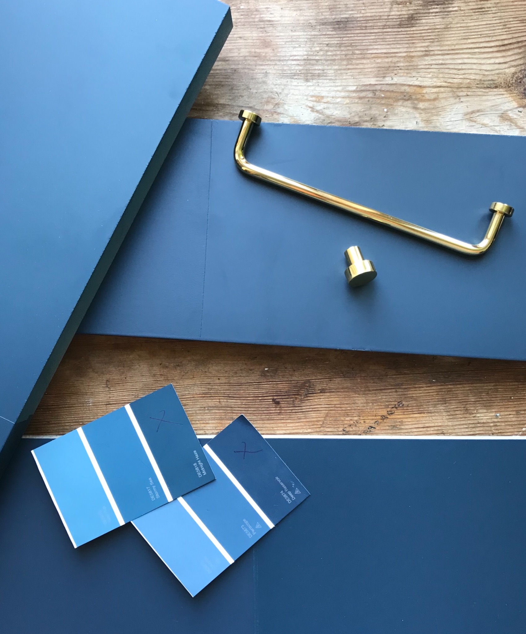 Niebieskie fronty meblowe z przymierzoną złotą gałką i uchwytem, dobrane próbki kolorów farb z zaznaczeniami na drewnianym blacie.