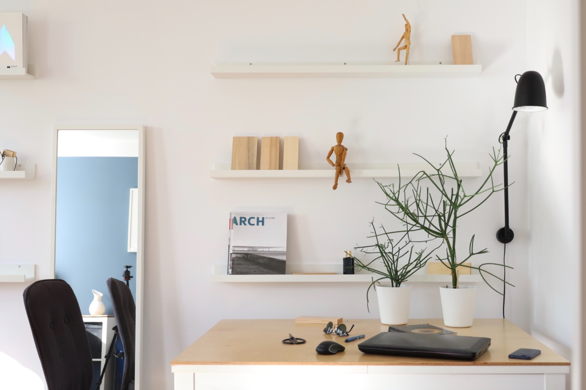 Minimalistyczne biuro domowe z białymi ścianami, drewnianym biurkiem, półkami z dekoracjami, lustrem i czarną lampą ścienną.