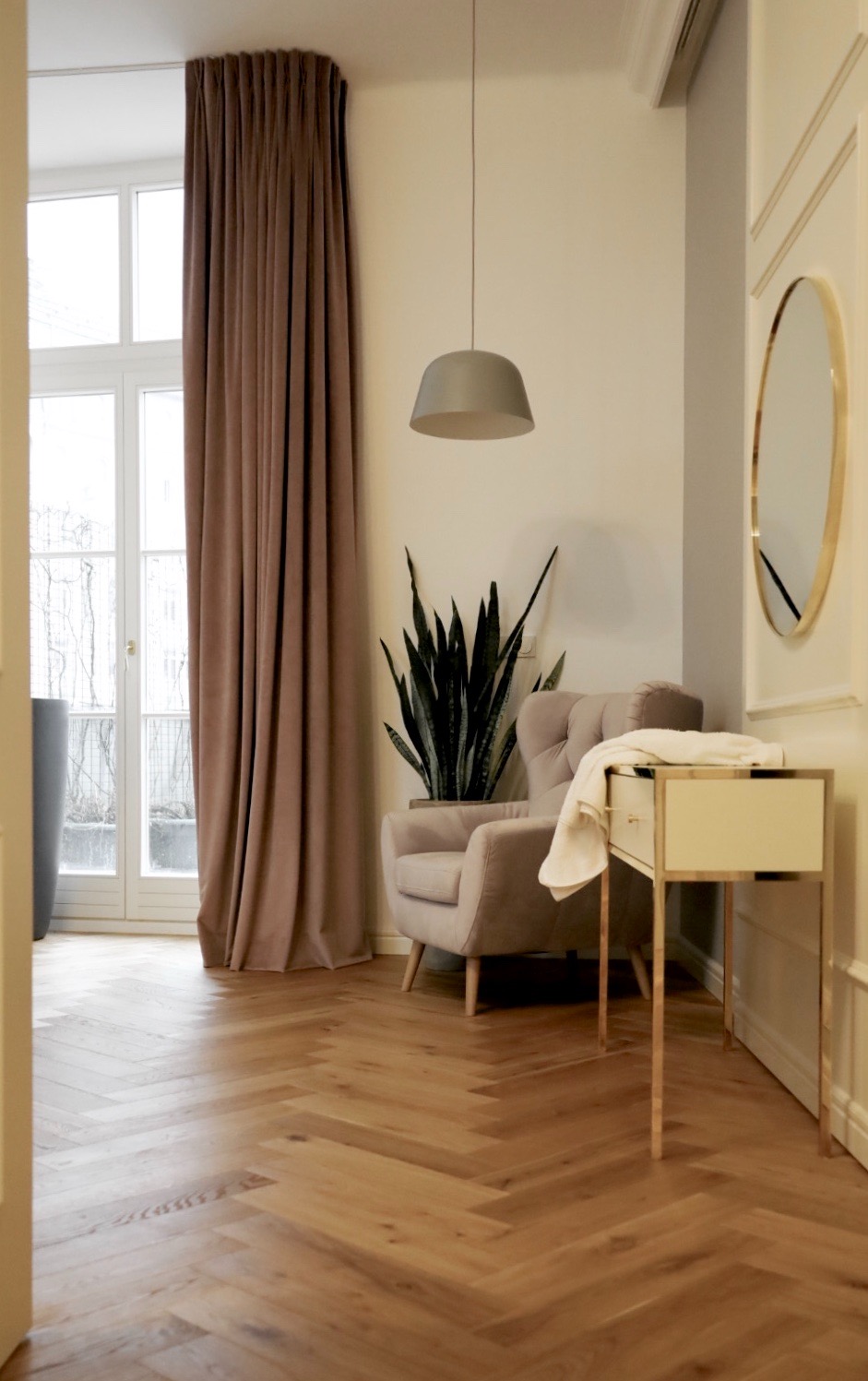 Jasny salon z parkietem ułożonym w jodełkę, beżowy fotel z rośliną, stolik z lustrem i brązowe zasłony przy oknie.