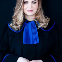 Portret młodej kobiety, prawnik, w todze adwokackiej z niebieskim żabotem, dłonie splecione, uśmiech na twarzy, na ciemnym tle.