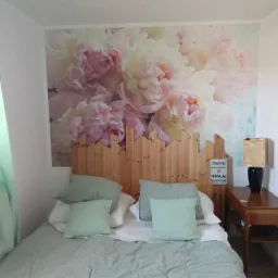 Jasna sypialnia z łóżkiem, drewnianym zagłówkiem z motywem palisady i fototapetą z kwiatami piwonii, uzupełniona miętową zasłoną i lampką nocną.