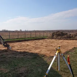 Przygotowanie terenu pod fundamenty: wykop z widocznym sprzętem geodezyjnym, mini koparką i stertą ziemi w tle, obok paleta z bloczkami fundamentowymi.