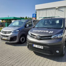 Dwa busy firmy transportowej z Rybnika, Opel i Toyota, zaparkowane na kostce brukowej przed myjnią bezdotykową. Na Toyocie widoczne logo firmy 'transportcik.pl'.