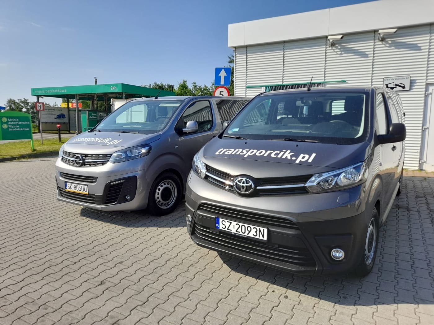 Dwa busy firmy transportowej z Rybnika, Opel i Toyota, zaparkowane na kostce brukowej przed myjnią bezdotykową. Na Toyocie widoczne logo firmy 'transportcik.pl'.