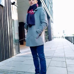 Młody mężczyzna w szarym płaszczu i granatowym szaliku, stojący na chodniku przed nowoczesnym budynkiem biurowym z przeszkloną fasadą i kamerą monitoringu.