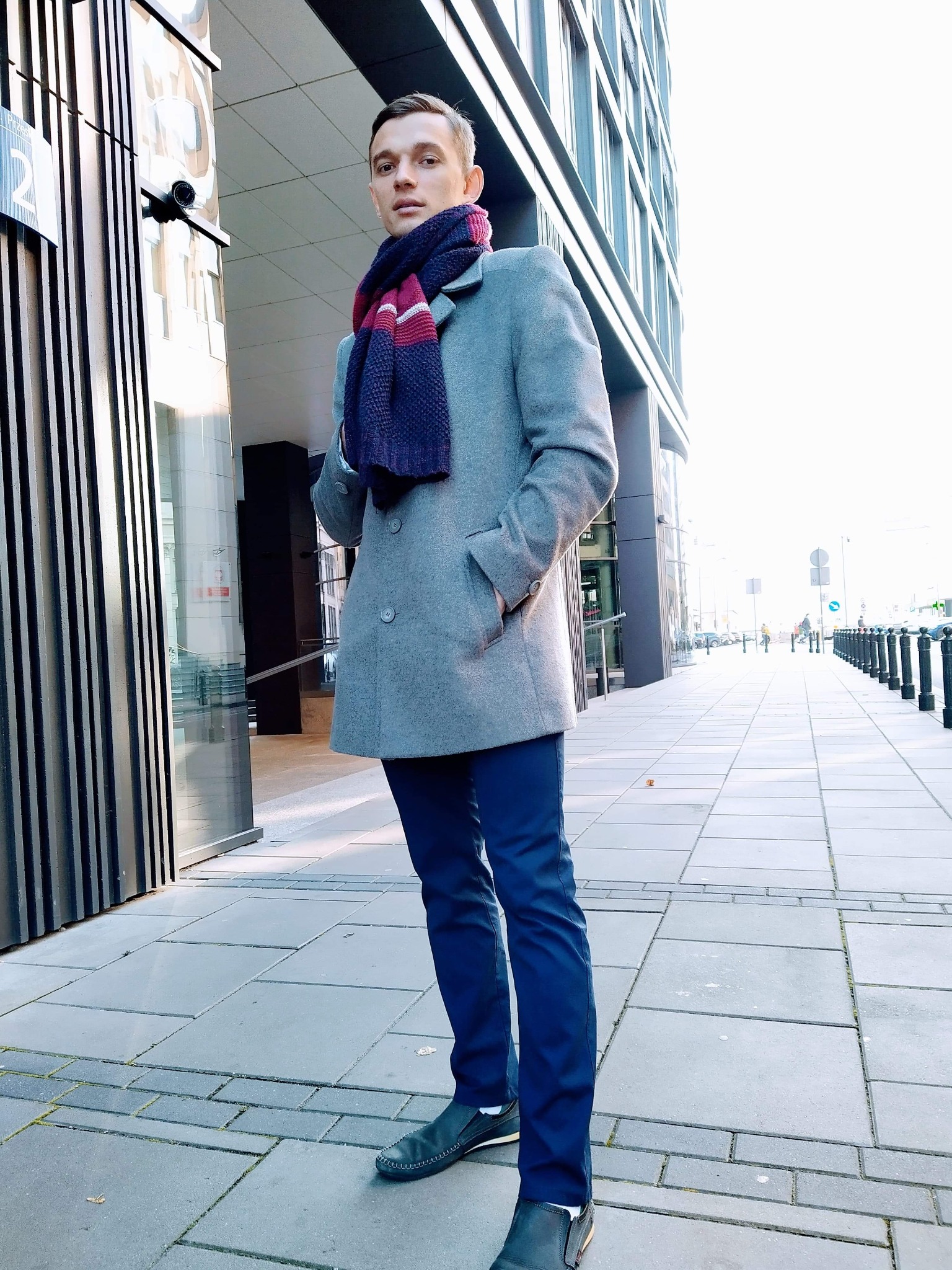 Młody mężczyzna w szarym płaszczu i granatowym szaliku, stojący na chodniku przed nowoczesnym budynkiem biurowym z przeszkloną fasadą i kamerą monitoringu.